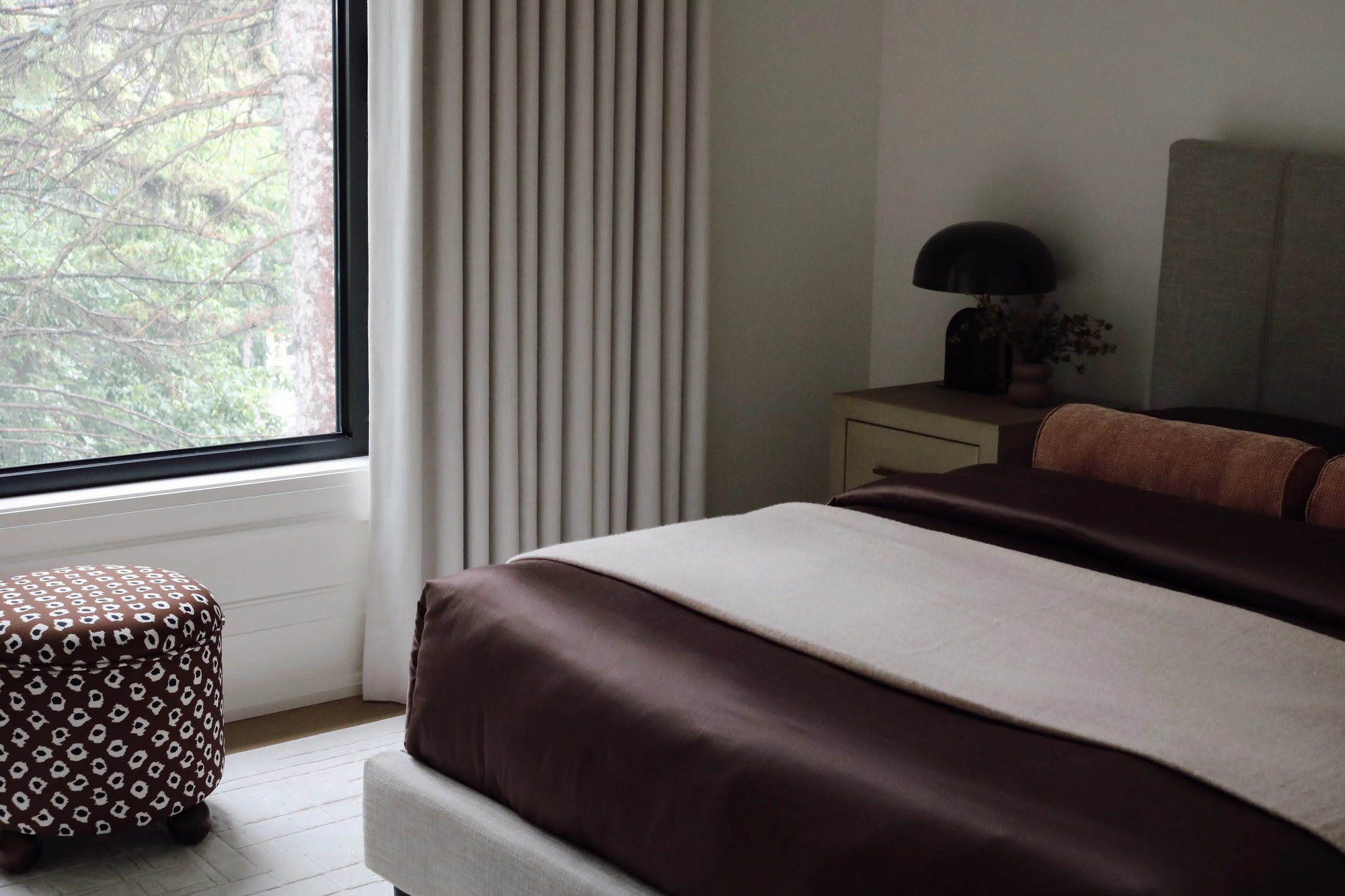 Floor to ceiling pleated custom drapery in a neutral fabric installed next to a large window in a modern bedroom with a dark wood bed, patterned ottoman, and warm lighting.